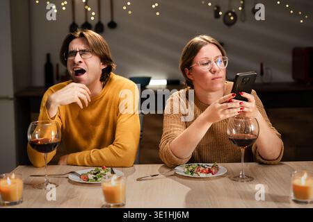 Loving couple having dinner with wine in kitchen at home. Wife scrolling smartphone while tired husband yawns after long working day. Evening family m Stock Photo