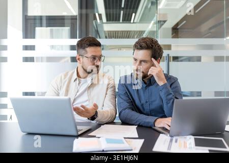 Two business colleagues sitting at a desk, looking at a laptop and working together, discussing ideas and collaborating on a project in a professional Stock Photo