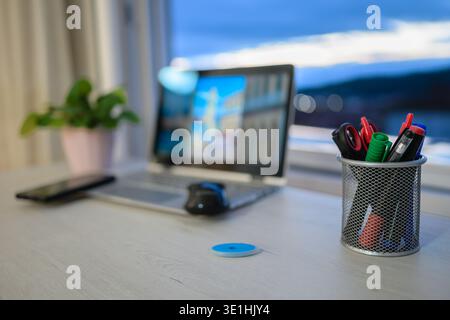 Modern home office workspace featuring a laptop, smartphone, and stationery, reflecting a contemporary remote work or study environment with evening l Stock Photo