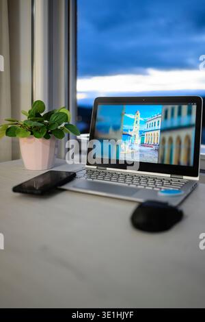 Modern laptop computer on a light desk, featuring a vibrant travel destination image on its screen, with a smartphone, mouse, and potted plant, beside Stock Photo