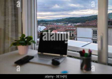 Modern laptop, smartphone, and plant sitting on a white desk in an indoor home office setup, looking out a window over a scenic town with traditional Stock Photo