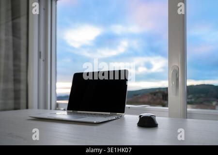 Laptop and mouse on a white desk by a large window with cloudy blue sky and distant town view, bright serene home office scene suggesting remote work Stock Photo