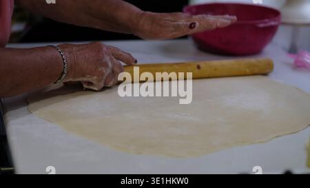 Hands roll out a thin sheet of dough using a wooden rolling pin on a kitchen work surface. The footage shows traditional home baking preparation Stock Photo