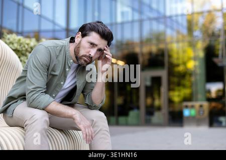 Stressed man feeling overwhelmed and having a headache, sitting on a modern city bench lost in thought, experiencing burnout and emotional stress outdoors in an urban area Stock Photo