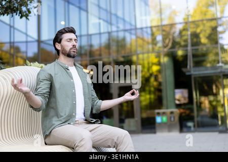 Man practicing mindfulness meditation in lotus position on a modern park bench, finding peace and balance amidst an urban building background, promoting mental wellness and healthy lifestyle Stock Photo