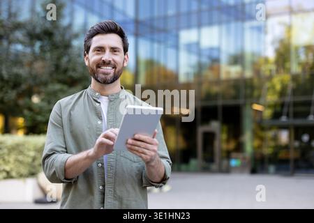 Smiling man feeling happy using a modern digital tablet for business communication, online learning, or casual browsing while standing outdoors in an urban setting Stock Photo