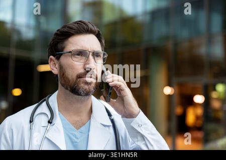 Male doctor in lab coat and stethoscope talks on smartphone outside a modern clinic, looking thoughtful and professional while offering telemedicine consultation and support Stock Photo