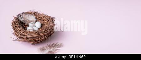 A natural nest containing several white eggs and decorative feathers, placed on a soft pink background, ideal for spring-themed designs Stock Photo