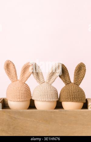 Three eggs wearing decorative crochet bunny hats in pink, beige, and brown, placed on a wooden surface with a soft pastel background Stock Photo