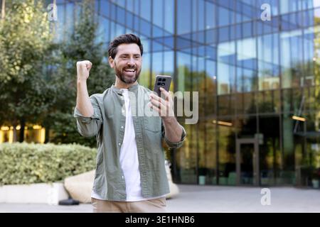 Excited businessman cheering and raising his fist while looking at a smartphone screen, standing outside a modern office building, celebrating a professional victory or good news Stock Photo