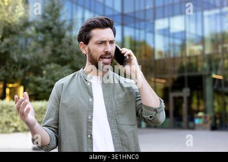 Man feeling frustration and anger during an unpleasant phone conversation, expressing disagreement and stress while communicating on a smartphone outdoors near a modern office building Stock Photo