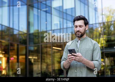 Young bearded man standing outdoors in front of a glass corporate building, focused on his smartphone for messaging, browsing and mobile connectivity in an urban business setting Stock Photo