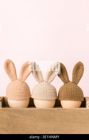 Three eggs wearing decorative crochet bunny hats in pink, beige, and brown, placed on a wooden surface with a soft pastel background Stock Photo