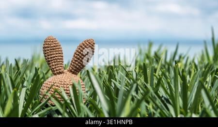 A crochet bunny with prominent ears is partially hidden in lush green grass, with a blurred ocean and sky in the background Stock Photo