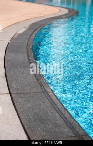 Curved stone border surrounds a swimming pool, showcasing clear blue water reflecting sunlight and creating a tranquil outdoor setting Stock Photo