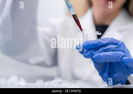 Automated pipetting system transferring red liquid or blood into microcentrifuge tubes at a clinical laboratory. Brightly lit workspace with various sample containers visible in the background. Stock Photo