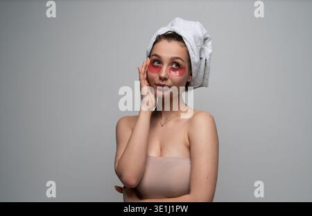 Young woman applying nourishing under eye patches and wearing a head towel, focusing on self-care and beauty routine for healthy skin Stock Photo