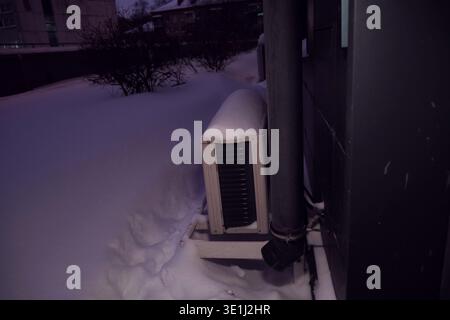 Snow covered air conditioning system installed on the exterior wall of a building during heavy winter snowfall. The quiet evening moment illustrates Stock Photo