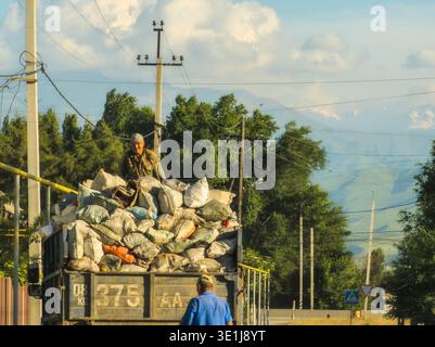 Garbage truck overloaded with sacks. Worker in camouflage stands atop load. Man in blue shirt nearby on dusty road in Gavrilovka, Chuy Province, Kyrgyzstan - June 4, 2024 Stock Photo