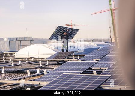 A worker carries a solar panel on a roof with lifting cranes in the background, PV installation at OBI Baumarkt, Muehlacker, Enzkreis, Germany Stock Photo
