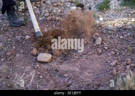 Worker using pickaxe to dig red soil, creating dust cloud, during outdoor work Stock Photo
