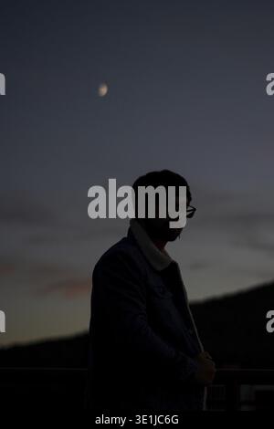 Silhouette of a young man observing the crescent moon in the twilight sky Stock Photo