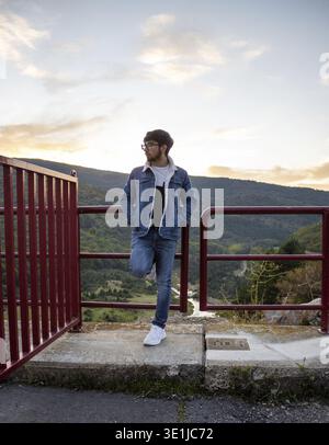 Thoughtful young man enjoying a beautiful sunset view in the mountains, leaning on a red railing Stock Photo
