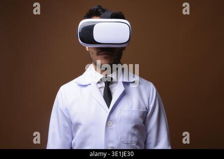 Studio shot of young handsome Hispanic man doctor using virtual reality headset against brown background Stock Photo