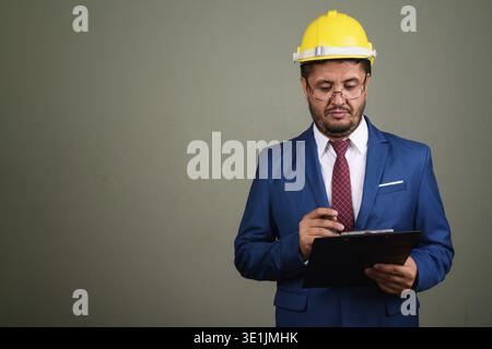 Studio shot of bearded Persian businessman wearing suit and hardhat against colored background Stock Photo