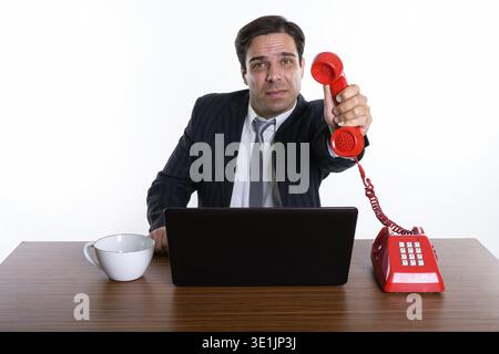 Studio shot of young Persian businessman giving old telephone with laptop and coffee cup on wooden table against white background Stock Photo