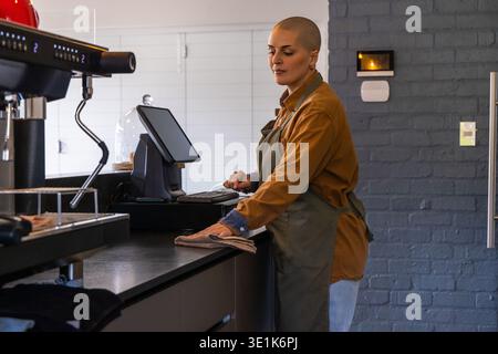 Female barista in olive apron at cafe using POS while wiping with cloth by espresso machine Stock Photo