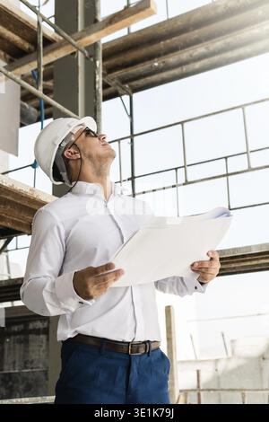 Young man architect or businessman wearing hard hat and holding blueprints on a construction site Stock Photo