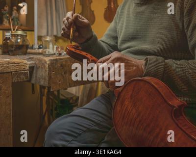 Close-up of a craftsman carefully working on a violin in a warm-toned workshop, Master violinmaker luthier hand varnishing the scroll of a new violin Stock Photo