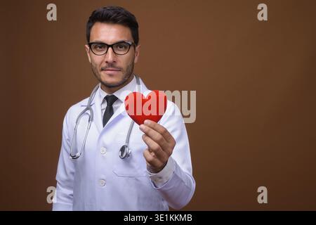 Studio shot of young handsome Hispanic man doctor wearing eyeglasses against brown background Stock Photo