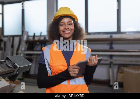 Portrait of a smiling African American female worker in a safety orange vest using a digital tablet in a metal processing factory. Stock Photo