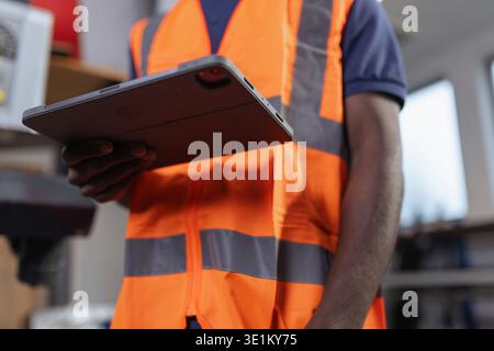 Close up of a smiling African American worker in a safety orange vest using a digital tablet in a metal processing factory. Stock Photo