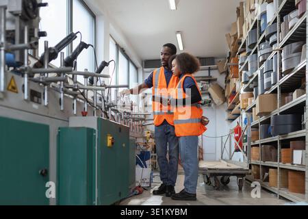 Technician african american workers wearing an orange safety vest using a tablet to control operating machinery at metal factory. Stock Photo
