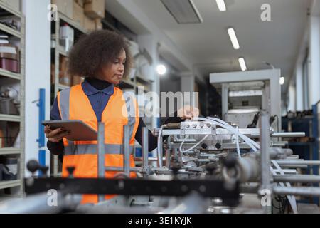 African American female worker operates in a modern, automated metal manufacturing facility, using a digital tablet for machinery diagnostics. Stock Photo