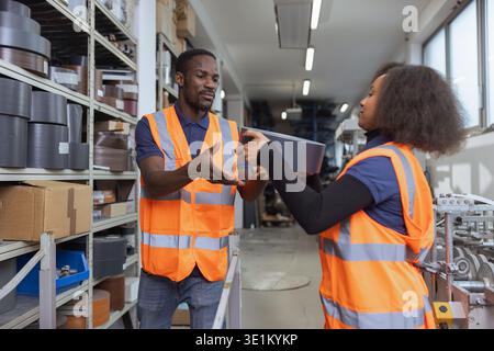 Two people, a male and a female warehouse worker collaborate in an industrial storage facility to safely lift and organise heavy inventory. Stock Photo