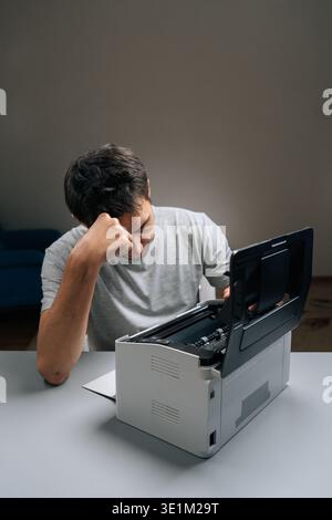 Frustrated man sitting at desk with paper jam in malfunctioning office printer, troubleshooting and trying to fix device amid stress and deadline Stock Photo