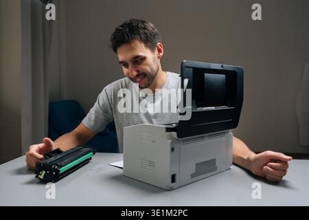 Positive repairman smiling performing printer maintenance, replacing empty toner cartridge with new one on desk, ensuring smooth office operations and Stock Photo
