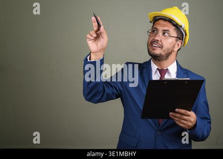 Studio shot of bearded Persian businessman wearing suit and hardhat against colored background Stock Photo