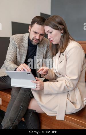 Colleagues holding and discussing tablet, focusing on work ideas while sitting close in indoor office. Tablet discussion, ideas, close sitting Stock Photo