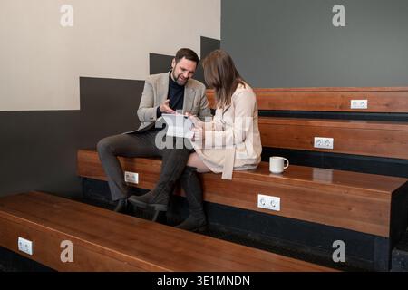 Male and female colleagues collaborating on business tasks on wooden bleachers in indoor workspace. Business collaboration, teamwork, workspace Stock Photo