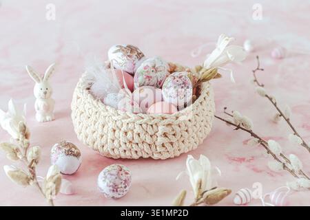 Easter decoration with pink and purple eggs with a bunny motif in a woven basket, decorated with magnolia flowers and willow branches, pink background Stock Photo