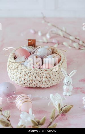 Easter decoration with pink and purple eggs with a bunny motif in a woven basket, decorated with magnolia flowers and willow branches, pink background Stock Photo