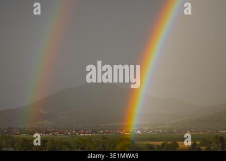Detailed view of a bright rainbow’s end touching a green valley landscape with a small town and mountains in the background. Stock Photo