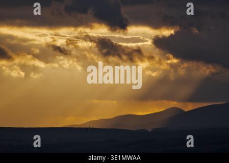 Intense crepuscular rays pierce through dark, heavy clouds during sunset, illuminating a silhouetted mountain range in a dramatic display of light and Stock Photo