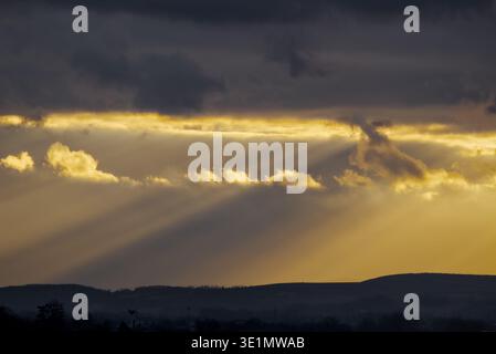 Dramatic crepuscular rays or "God rays" shining through a gap in dark, heavy clouds over a dark silhouetted mountain horizon. Stock Photo