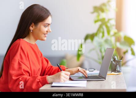 Woman Working on Laptop While Taking Notes at Desk in Bright Room During Daytime Stock Photo